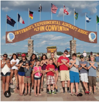 A group of children from the Bonanza group gather in front of the EAA sign , on the Air show grounds. They have all been coming here for multiple years, most of them, including Evans, since they were just a year old. They take a photo in front of this sign every year to commemorate the adventures they have in Oshkosh. In this photo, taken in 2015, sophomore Sydney Evans and junior Lauryn Kylop, Grace Matther, and Sarita Evans are all standing on the right. 